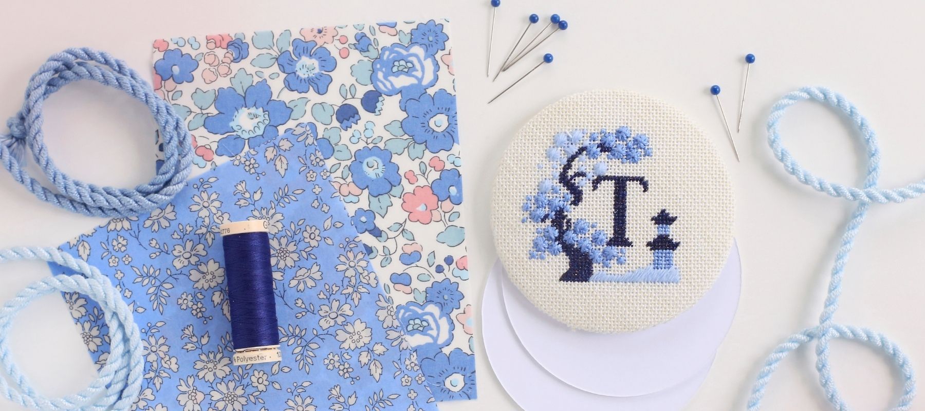 Chinoiserie - Three Charted Monograms project with the letter T, surrounded by blue cording, blue thread, blue floral finishing fabric, and pins.