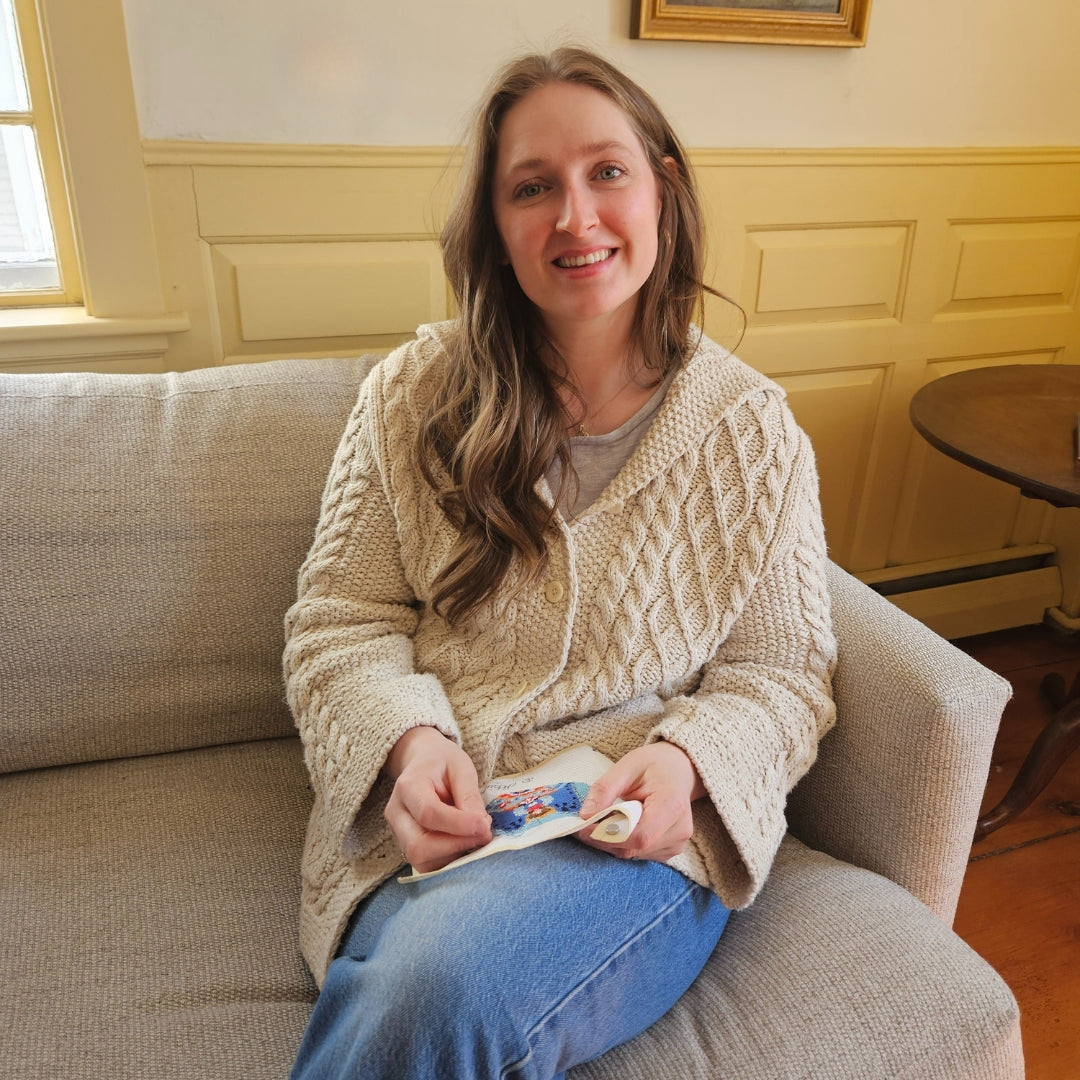 A woman with a cream sweater is sitting on a sofa working on a needlepoint project.