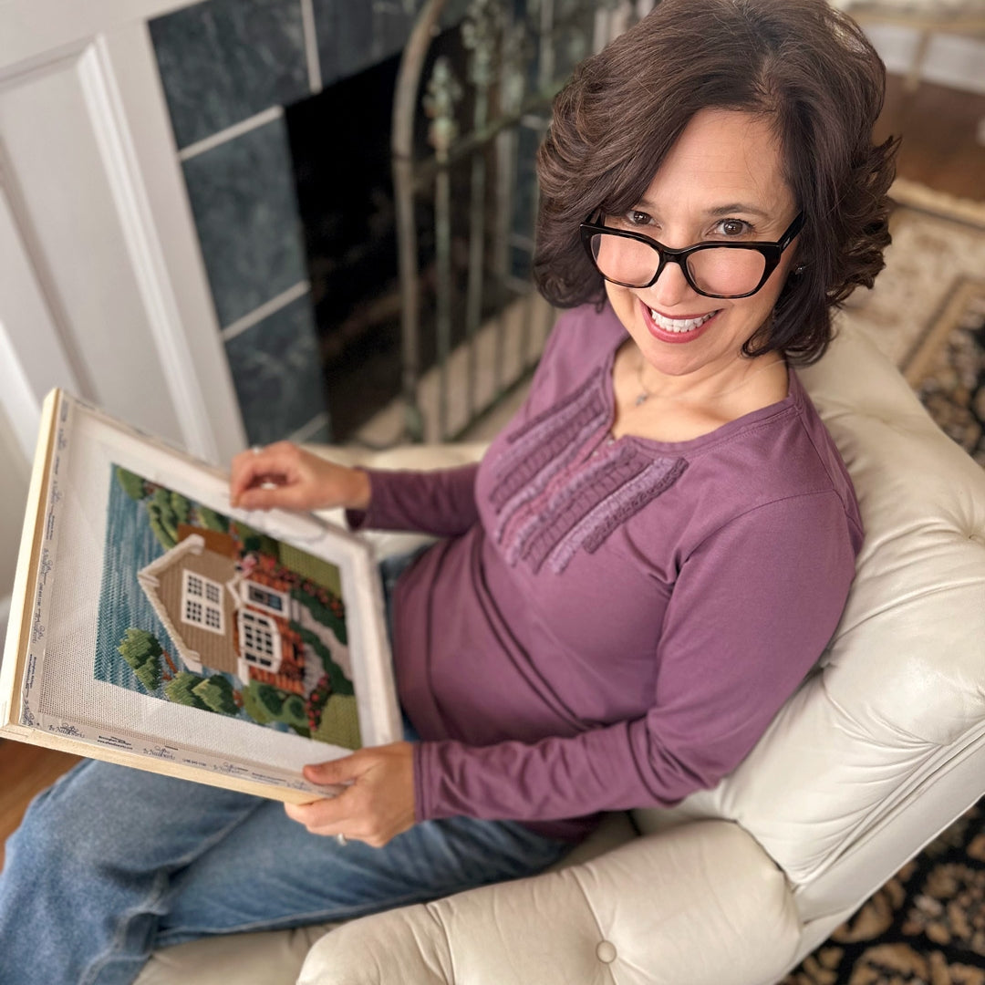 A woman is looking at a completed, framed needlepoint house portrait while sitting on a loveseat.