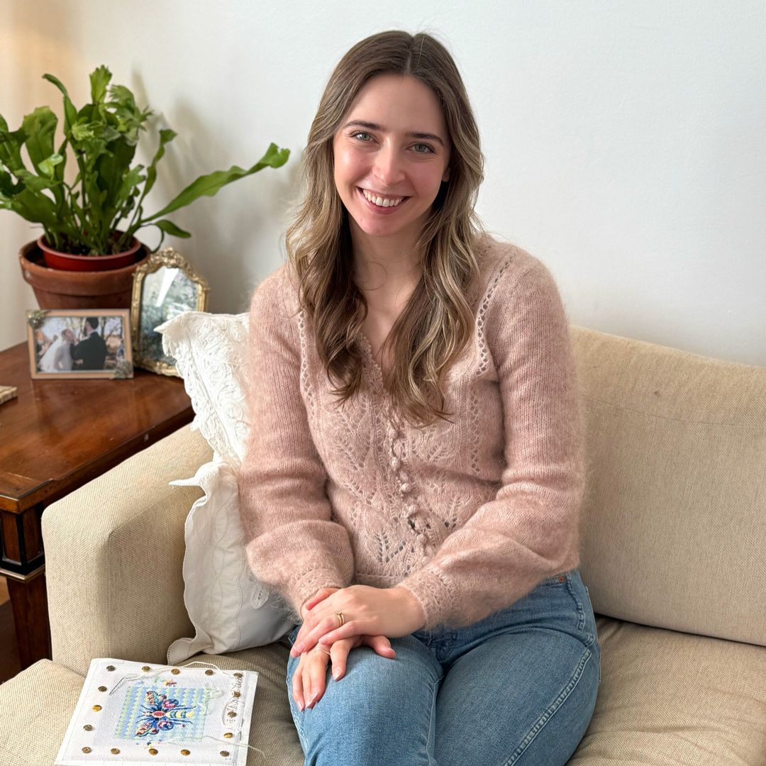 A woman is sitting on a couch next to a butterfly needlepoint project smiling at the camera.
