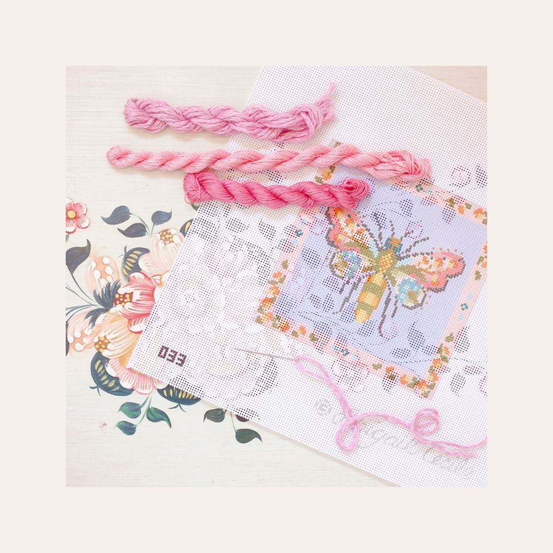 A painted needlepoint canvas of a colorful bee, surrounded by thread in different shades of pink. 