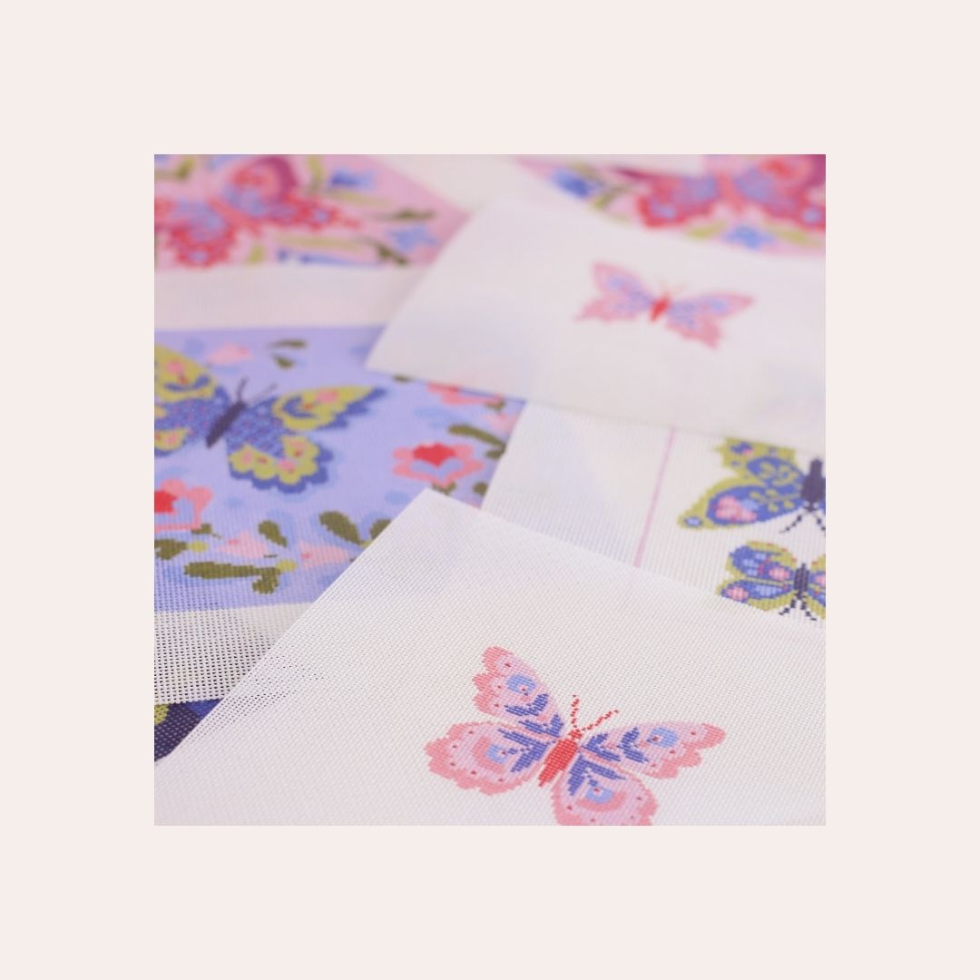 Various butterfly painted needlepoint canvases in shades of pink and purple spread across a tabletop.
