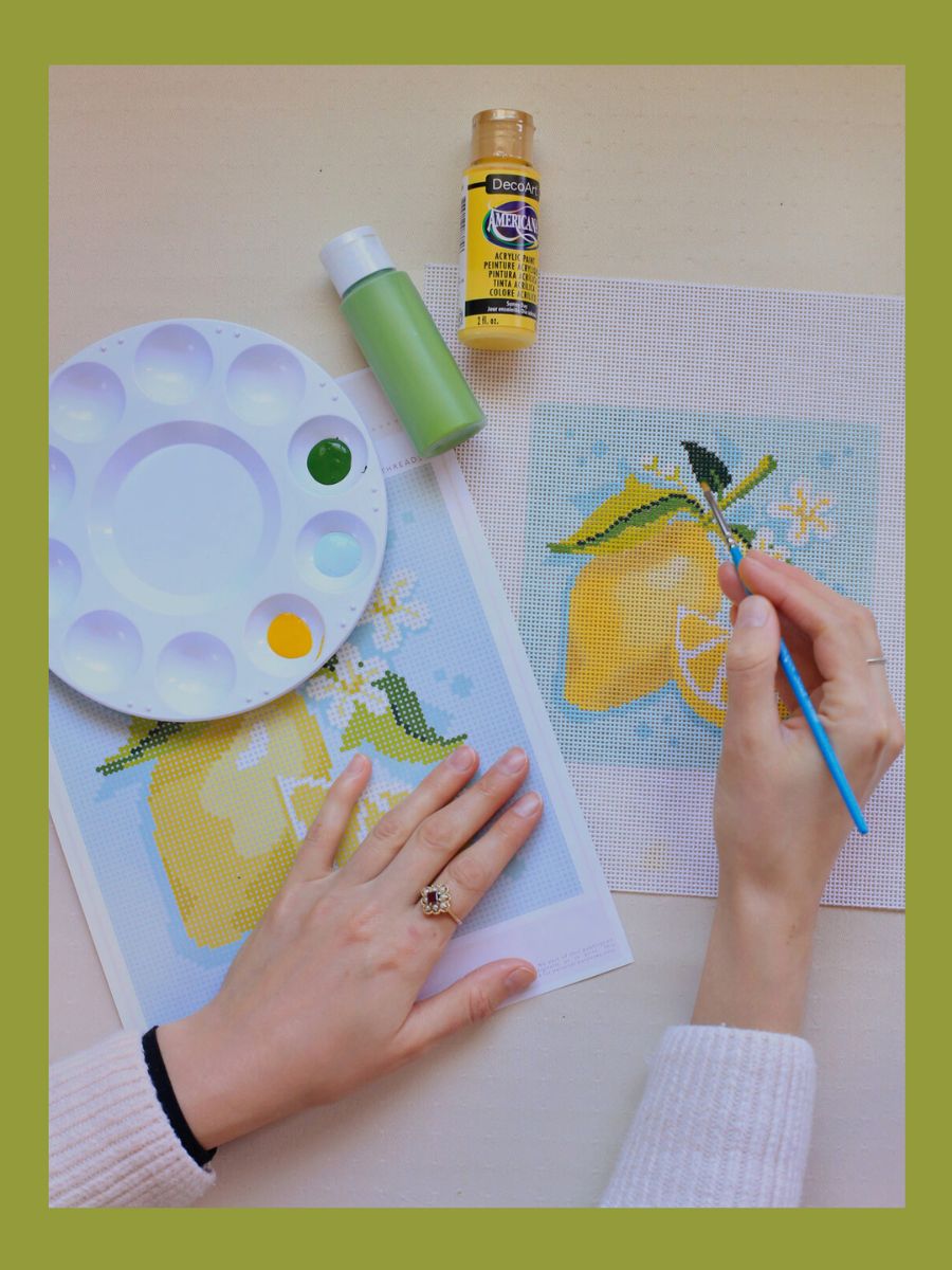 A woman is painting a lemon on a needlepoint canvas.
