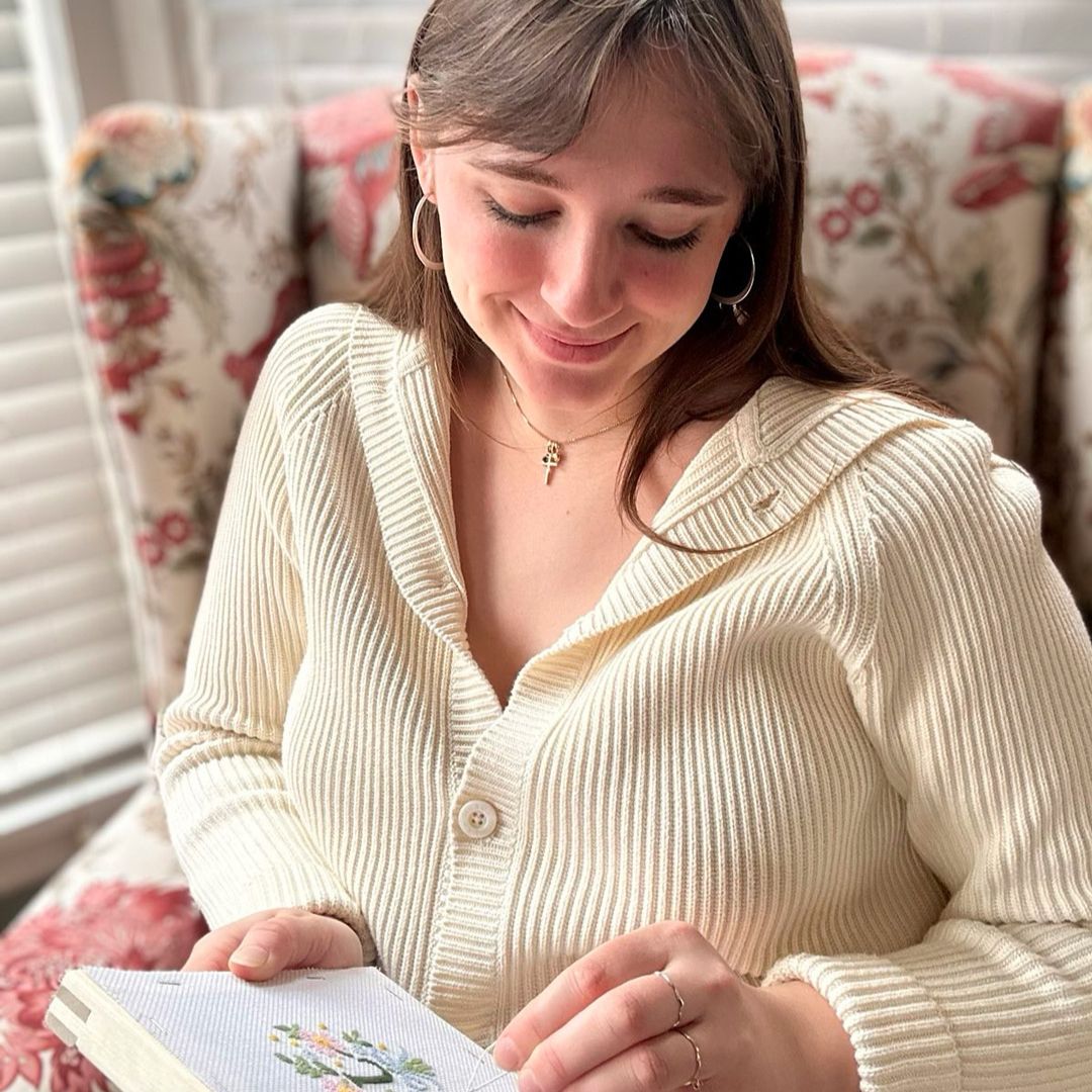 A woman wearing a cream cardigan is stitching a monogram needlepoint while sitting on a tapestry chair.