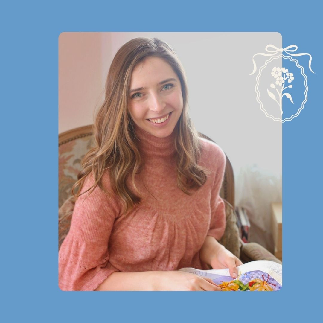 A woman is smiling while sitting on a chair working on a needlepoint project.