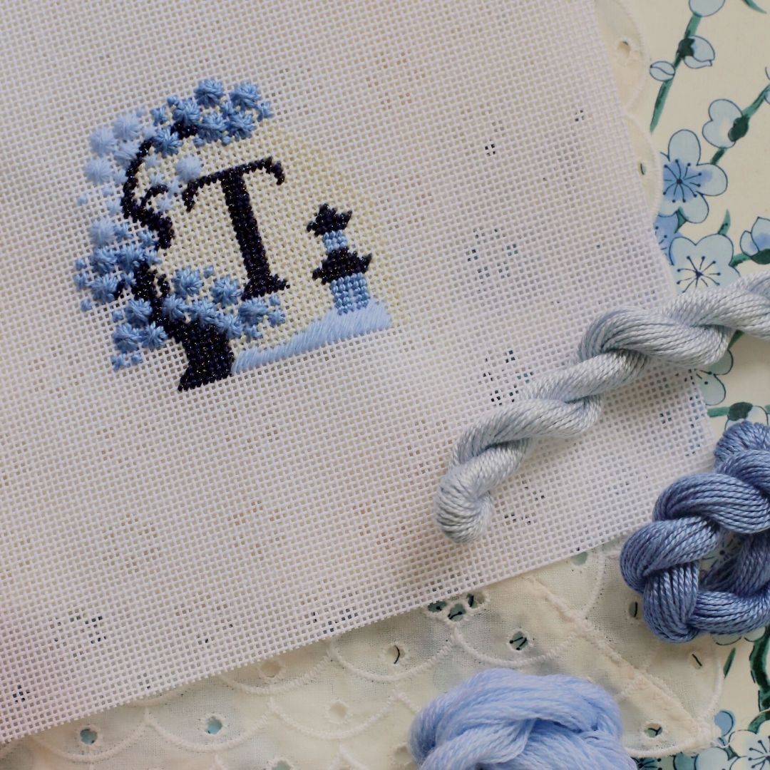A Chinoiserie - Three Charted Monogram project with the letter T is almost completed. It sits on a table top surrounded by thread in different shades of blue.
