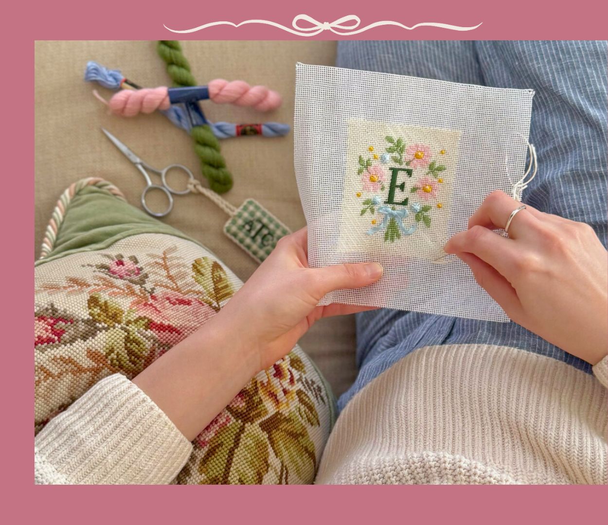 A woman is stitching a monogram needlepoint project while sitting on a sofa.