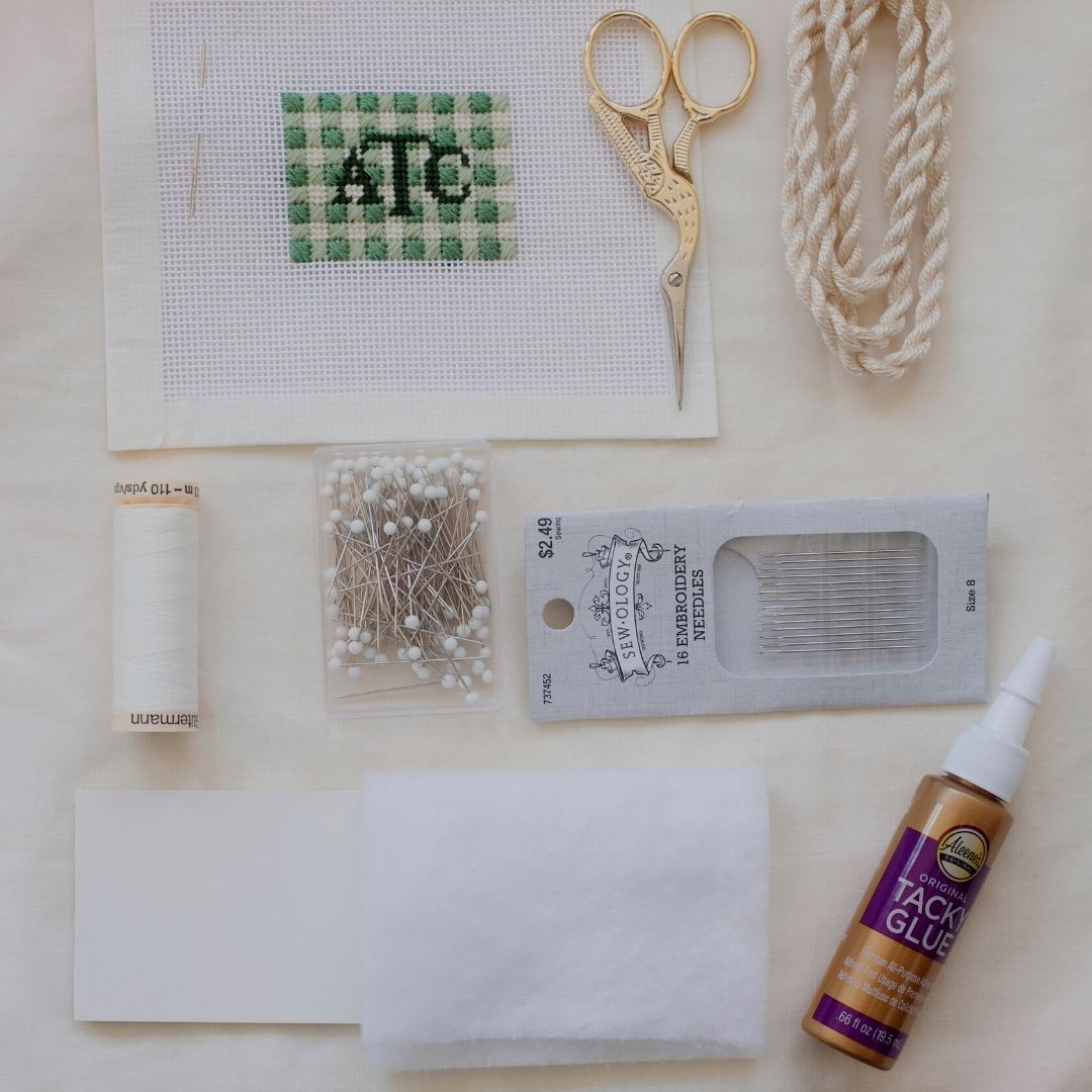 Monogram scissor fob project with the monograms "ATC", surrounded by needlepoint tools - a pair of scissors, cording, thread, box of pins, pack of needles, and tacky glue.