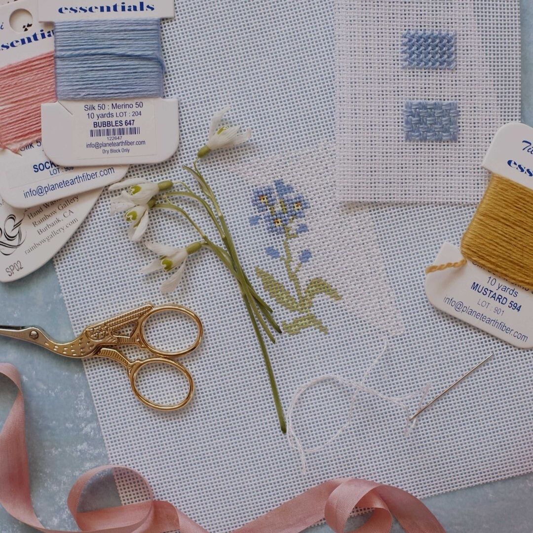 Blue wildflower needlepoint project in process, surrounded by needlepoint tools including a pair of gold scissors, and pink, blue and gold thread.