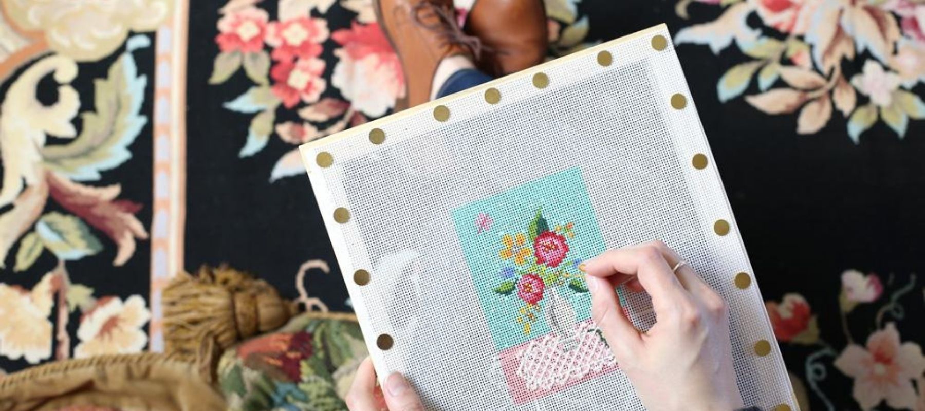 Woman is sitting in her chair, needlepointing a scene with colorful flowers in a vase. 