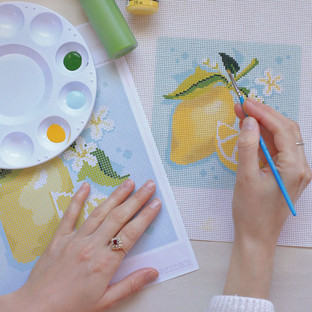 A woman is painting a lemon on needlepoint canvas.