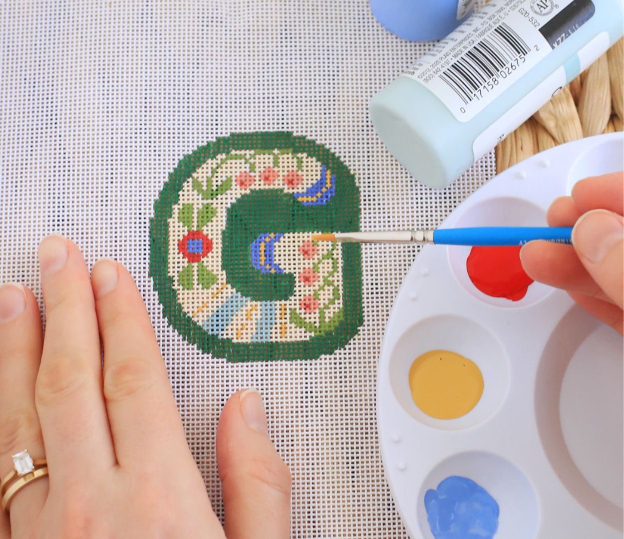 A woman is painting a monogram on a needlepoint canvas.