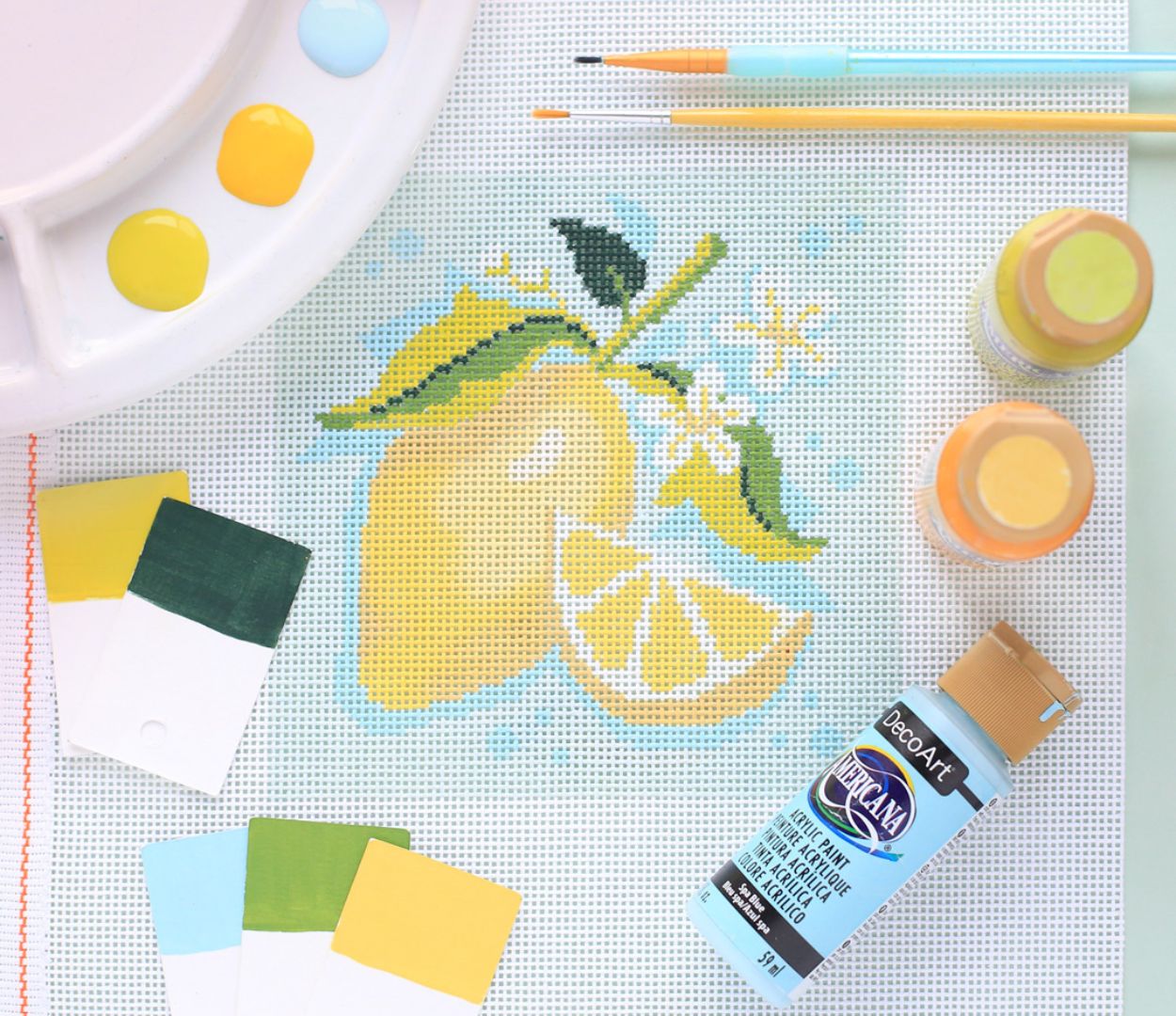 A lemon painted on a needlepoint canvas is laying next to a paint bottle, paint swatches, brushes, and a palette.