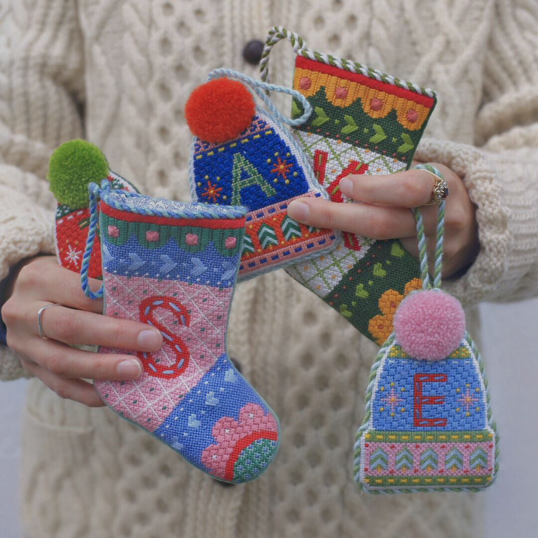 A woman in a white sweater is holding several completed holiday needlepoint ornaments.