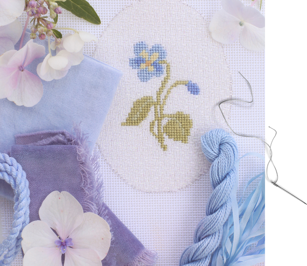 Blue wildflower needlepoint project with completed stitching, on a tabletop surrounded by fresh flowers, finishing fabric, thread, and cording.
